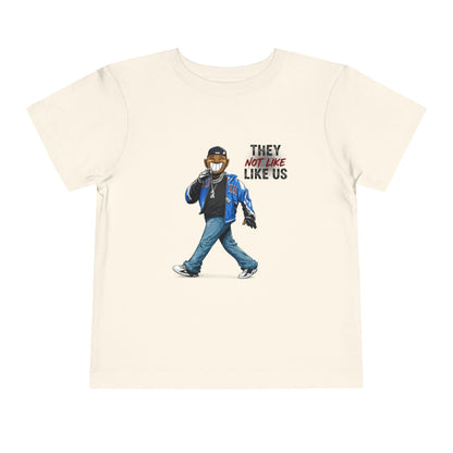 Beige t-shirt with graphic of a person wearing a mask and text 'They Not Like Us' on a white background