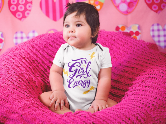 Baby wearing a 'Girl Energy' onesie sitting on a pink textured surface with a colorful heart-patterned background.